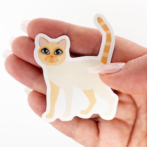 May include: A sticker of a Siamese cat with a cream-colored body and tan markings on its ears, paws, and tail. The cat has blue eyes and a detailed face. The sticker is held in a person's hand against a white background.
