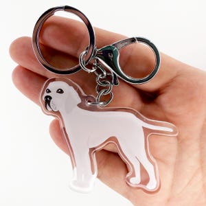 May include: A clear acrylic keychain featuring a white dog design with black accents. The keychain has a silver metal ring and clasp for attaching to keys or bags. The dog is standing, with its features clearly defined.
