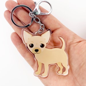 May include: A keychain featuring a cartoon chihuahua dog in tan and white. The dog has large black eyes and is attached to a silver keyring and clasp. The keychain is made of clear acrylic.