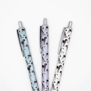May include: Three ballpoint pens with silver accents and a repeating pattern of brown dogs and bones. The pens have a light blue, lavender, and white background. The pens are angled towards the center.