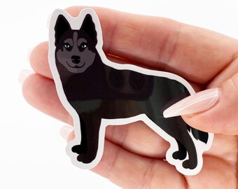 May include: A black and white sticker featuring a stylized illustration of a husky dog. The dog has green eyes and a dark gray face. The sticker is held in a hand against a white background.