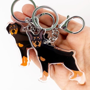 May include: Two acrylic keyrings with a black and tan Rottweiler dog design. The keyrings have silver-coloured metal hardware, including a split ring and a lobster clasp. The dog's features are detailed with orange and brown accents.