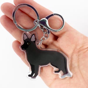 May include: A black German Shepherd dog keychain with a silver keyring and clasp. The dog is depicted in a standing pose with a black body, gray ears, and a black nose. The keychain is made of clear acrylic.