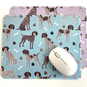 May include: A blue and white mouse pad with a repeating pattern of brown and white German Shorthaired Pointers. The mouse pad is layered on top of a purple and white mouse pad with the same pattern. A white computer mouse is on top of the blue and white mouse pad.