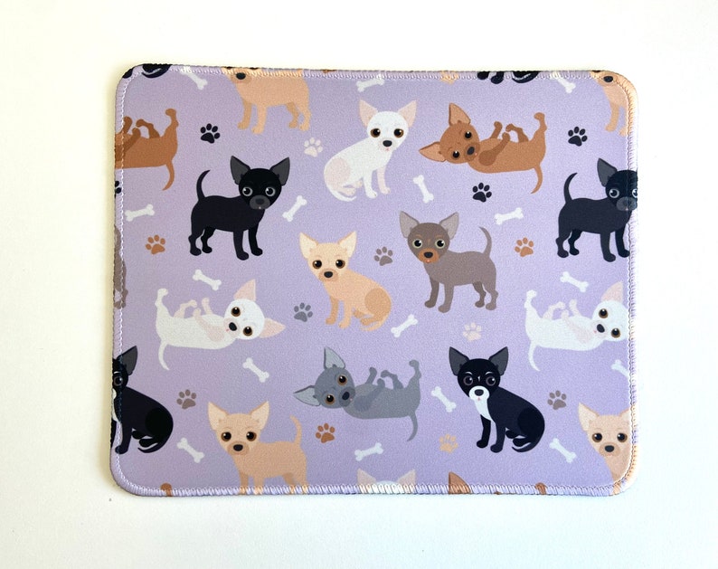 May include: A purple mouse pad with a repeating pattern of cartoon chihuahua dogs in various colors and poses. The dogs are on a light purple background with white paw prints.