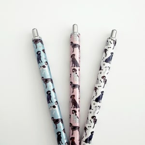 May include: Three pens with dog patterns. One pen is light blue with black and white dogs, another is pink with brown and white dogs, and the third is white with black dogs and bone accents. The pens have silver tips.