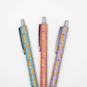 May include: Three pens with silver clips and tips. The pens have a repeating pattern of orange tabby cats on different colored backgrounds: teal, pink, and purple. The pens are arranged in a fan shape against a white background.