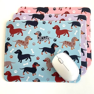 May include: A set of four mousepads featuring a repeating pattern of cartoon dachshunds in various colors and poses. The mousepads are layered on top of each other, with the top mousepad being light blue with white bone-shaped accents. The mousepads are made of a soft, fabric material.