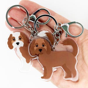 May include: Two dog-themed keychains with silver-colored hardware. One keychain features a brown and white Cavalier King Charles Spaniel design, and the other has a brown dog design. Both keychains have a clear, glossy finish.