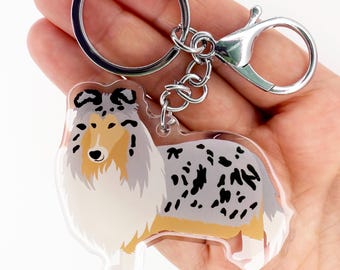 May include: A clear acrylic keychain featuring a stylized illustration of a Collie dog. The dog has a blue merle coat with black spots, a white chest, and tan markings. The keychain has a silver keyring and clasp.