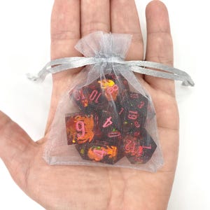 Op de afbeelding: Een set polyhedrale dobbelstenen in een doorzichtig, zilverkleurig zakje met trekkoord. De dobbelstenen zijn donker met rode en oranje accenten en roze cijfers. De tas wordt in een open hand tegen een witte achtergrond gehouden.