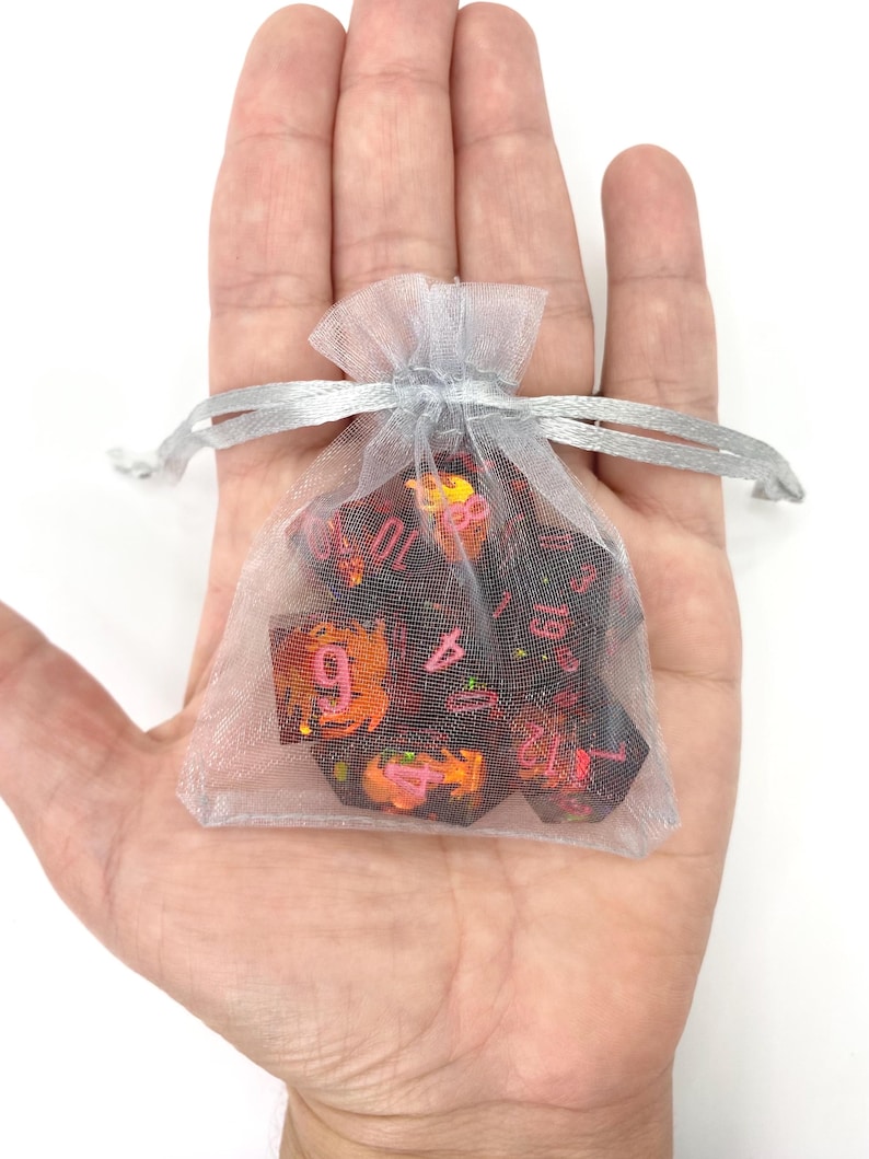 Op de afbeelding: Een set polyhedrale dobbelstenen in een doorzichtig, zilverkleurig zakje met trekkoord. De dobbelstenen zijn donker met rode en oranje accenten en roze cijfers. De tas wordt in een open hand tegen een witte achtergrond gehouden.