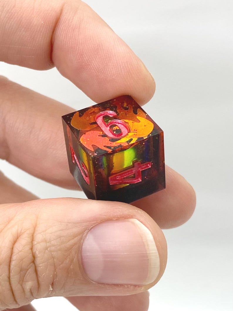 May include: A square, multi-sided die with red numerals and a fiery, orange and yellow interior. The die is held between two fingers, showcasing its intricate design and color scheme. The die is a gaming accessory.