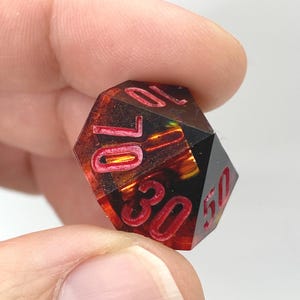 Op de afbeelding: Een veelzijdige, rood-zwarte dobbelsteen met witte cijfers. De dobbelsteen wordt tussen de vingers gehouden en heeft de cijfers 10, 30 en 50. De dobbelsteen heeft een vurige, doorschijnende binnenkant.