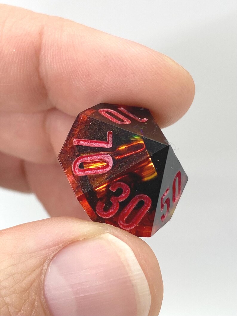 Op de afbeelding: Een veelzijdige, rood-zwarte dobbelsteen met witte cijfers. De dobbelsteen wordt tussen de vingers gehouden en heeft de cijfers 10, 30 en 50. De dobbelsteen heeft een vurige, doorschijnende binnenkant.