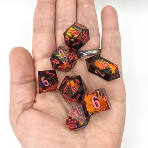 Op de afbeelding: Een verzameling van zeven polyhedrale dobbelstenen met een oranje en zwarte kleurstelling en roze cijfers. De dobbelstenen zijn ontworpen voor tabletop rollenspellen. De dobbelstenen worden in een hand gehouden.