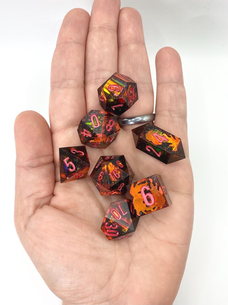 Op de afbeelding: Een verzameling van zeven polyhedrale dobbelstenen met een oranje en zwarte kleurstelling en roze cijfers. De dobbelstenen zijn ontworpen voor tabletop rollenspellen. De dobbelstenen worden in een hand gehouden.