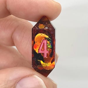 Op de afbeelding: Een handgemaakte, facetgeslepen hars hanger met het roze nummer 4. De hanger is gevuld met oranje, gouden en iriserende vlokken. De hars heeft een diepe amberkleur, vastgehouden tussen vingers.