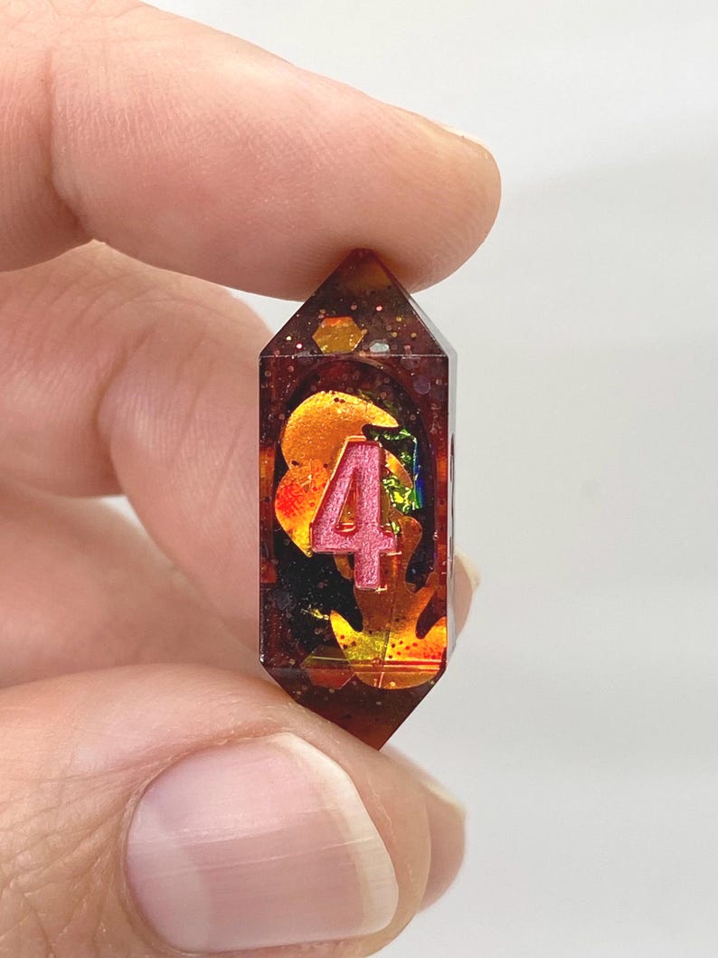 Op de afbeelding: Een handgemaakte, facetgeslepen hars hanger met het roze nummer 4. De hanger is gevuld met oranje, gouden en iriserende vlokken. De hars heeft een diepe amberkleur, vastgehouden tussen vingers.