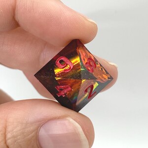 Op de afbeelding: Een veelzijdige D20 dobbelsteen, die op een edelsteen lijkt. De dobbelsteen is een mix van amber, oranje en bruin met rode cijfers. De dobbelsteen wordt tussen twee vingers tegen een witte achtergrond gehouden. De dobbelsteen is voor tabletop games.