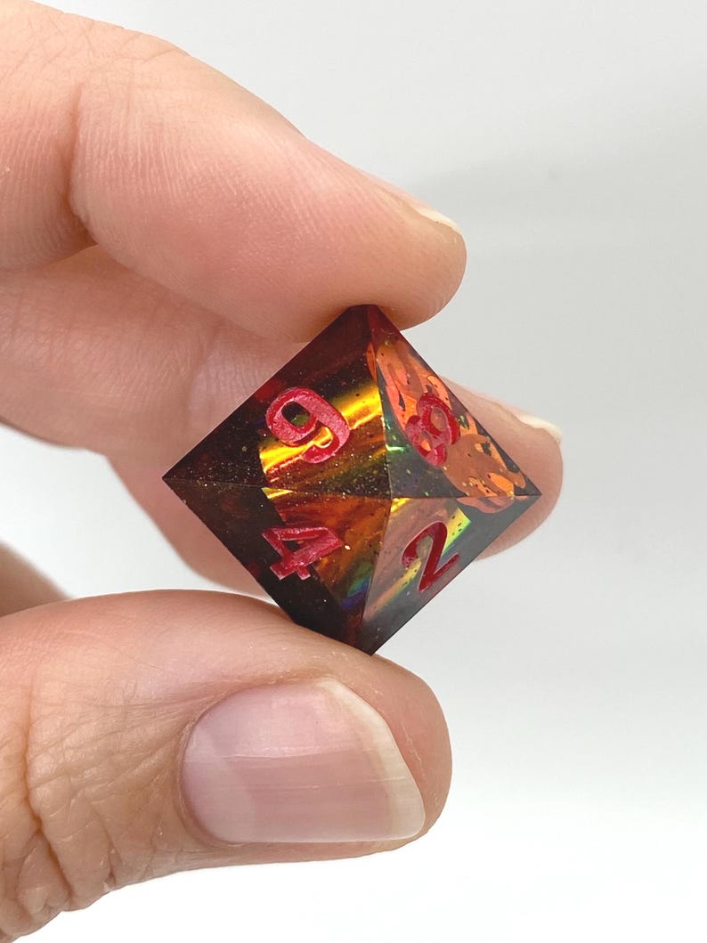 Op de afbeelding: Een veelzijdige D20 dobbelsteen, die op een edelsteen lijkt. De dobbelsteen is een mix van amber, oranje en bruin met rode cijfers. De dobbelsteen wordt tussen twee vingers tegen een witte achtergrond gehouden. De dobbelsteen is voor tabletop games.