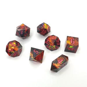 Op de afbeelding: Set van zeven polyhedrale dobbelstenen met een donkere basis en levendige rode en oranje interne ontwerpen. De cijfers zijn felroze van kleur. Deze dobbelstenen zijn ontworpen voor tabletop-rollenspellen.