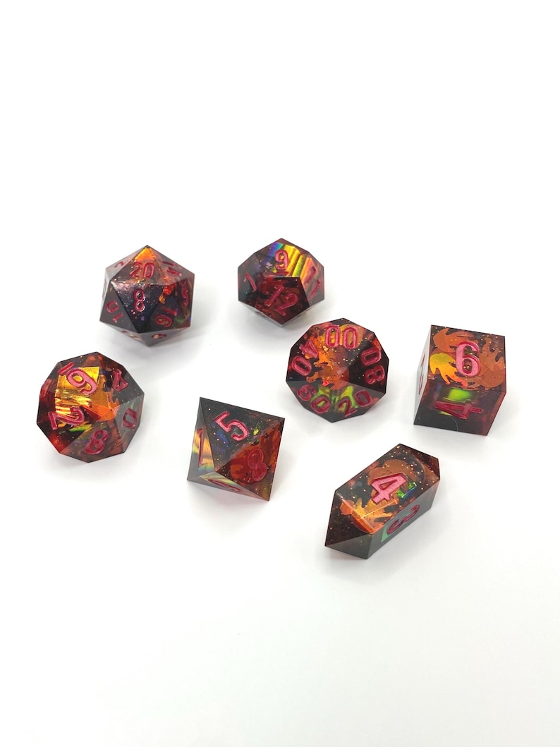 Op de afbeelding: Set van zeven polyhedrale dobbelstenen met een donkere basis en levendige rode en oranje interne ontwerpen. De cijfers zijn felroze van kleur. Deze dobbelstenen zijn ontworpen voor tabletop-rollenspellen.