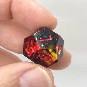 Op de afbeelding: Een veelzijdige dobbelsteen met rode cijfers, die op een edelsteen lijkt. De dobbelsteen is een mix van rood, zwart en goud, met glitteraccenten. Het nummer 1 is zichtbaar op de bovenkant. De dobbelsteen wordt tussen twee vingers gehouden.