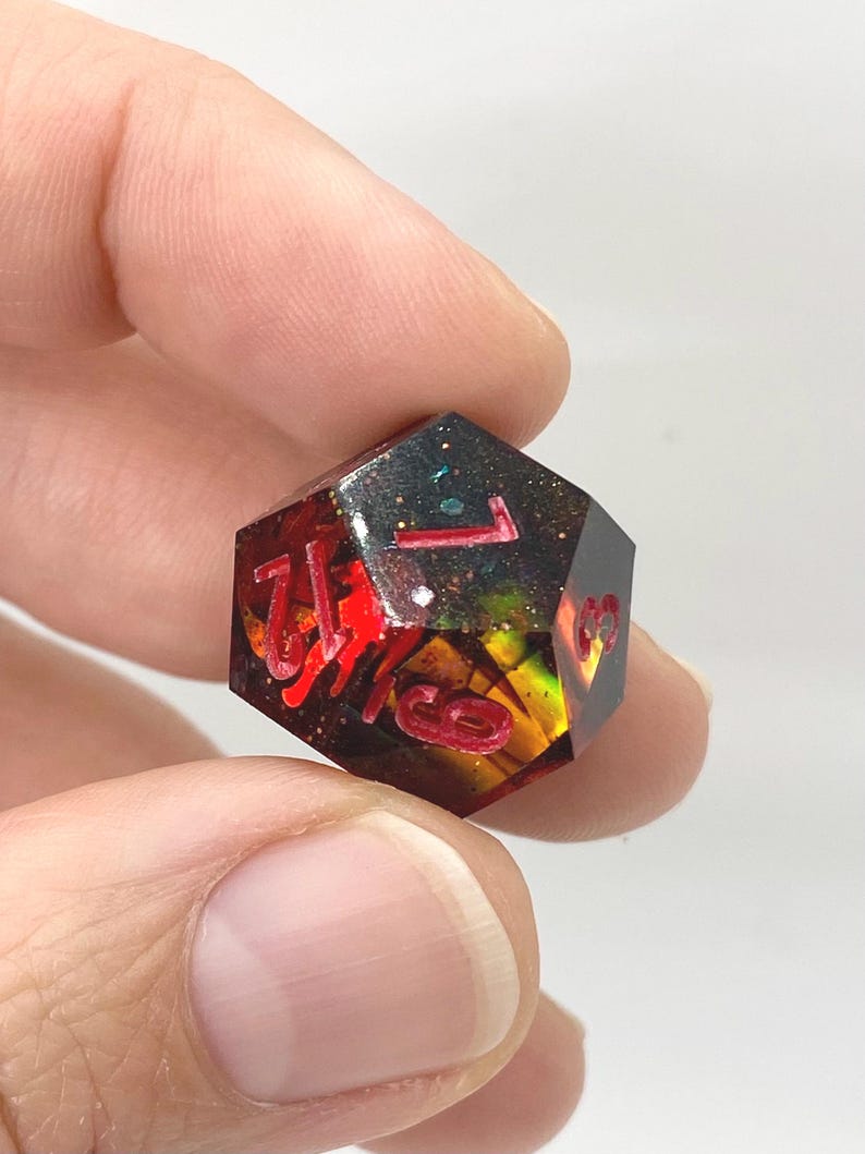 Op de afbeelding: Een veelzijdige dobbelsteen met rode cijfers, die op een edelsteen lijkt. De dobbelsteen is een mix van rood, zwart en goud, met glitteraccenten. Het nummer 1 is zichtbaar op de bovenkant. De dobbelsteen wordt tussen twee vingers gehouden.