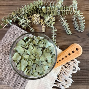 May include: A clear glass bowl filled with dried green eucalyptus leaves, positioned on a textured white and brown fabric with fringe. A light-colored wooden tool with a series of circular holes lies beside the bowl, accompanied by sprigs of eucalyptus.