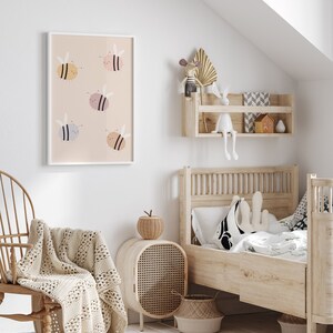 Puede incluir: Una impresi&oacute;n enmarcada en blanco con un fondo rosa claro y seis abejas de dibujos animados en varios tonos de rosa, amarillo y azul. La impresi&oacute;n est&aacute; colgada en una pared blanca en la habitaci&oacute;n de un ni&ntilde;o. La habitaci&oacute;n tambi&eacute;n cuenta con una cama de madera con una almohada con estampado blanco y negro, una mesita de noche de mimbre y una manta tejida en una mecedora.