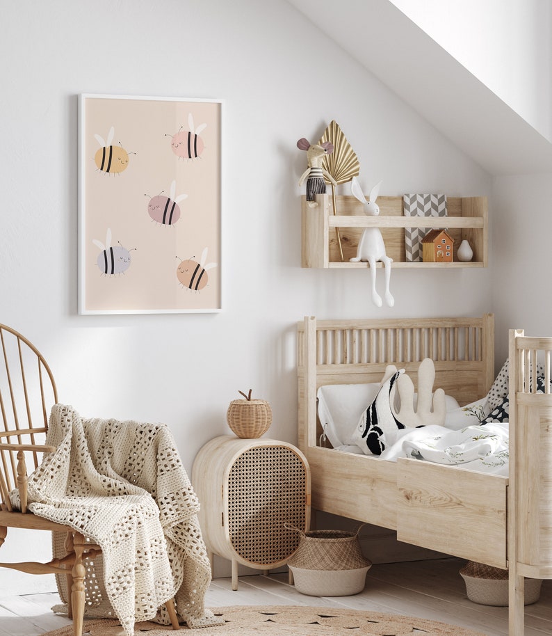 Puede incluir: Una impresi&oacute;n enmarcada en blanco con un fondo rosa claro y seis abejas de dibujos animados en varios tonos de rosa, amarillo y azul. La impresi&oacute;n est&aacute; colgada en una pared blanca en la habitaci&oacute;n de un ni&ntilde;o. La habitaci&oacute;n tambi&eacute;n cuenta con una cama de madera con una almohada con estampado blanco y negro, una mesita de noche de mimbre y una manta tejida en una mecedora.