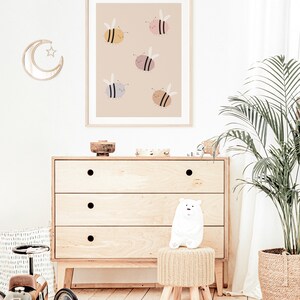 Puede incluir: Una impresi&oacute;n enmarcada beige con un fondo marr&oacute;n claro que presenta cinco abejas de dibujos animados en varios colores, incluyendo amarillo, rosa, gris y marr&oacute;n. Las abejas est&aacute;n volando y tienen expresiones felices.