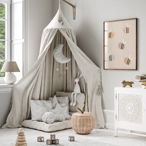 Puede incluir: Una cama con dosel blanco con un colch&oacute;n y almohadas blancas mullidas. Un conejito de peluche est&aacute; sentado en la cama, y una cesta de mimbre con una manzana de tela en la parte superior est&aacute; en el suelo. Hay bloques de madera con letras en ellos en el suelo. Una impresi&oacute;n enmarcada con un fondo rosa claro y abejas coloridas cuelga de la pared.