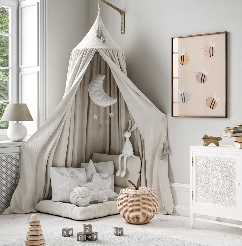 Puede incluir: Una cama con dosel blanco con un colch&oacute;n y almohadas blancas mullidas. Un conejito de peluche est&aacute; sentado en la cama, y una cesta de mimbre con una manzana de tela en la parte superior est&aacute; en el suelo. Hay bloques de madera con letras en ellos en el suelo. Una impresi&oacute;n enmarcada con un fondo rosa claro y abejas coloridas cuelga de la pared.