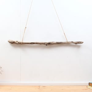 Op de afbeelding: Een natuurlijke drijfhouten plank, opgehangen met touw tegen een witte muur. De plank is lichtbruin van kleur met een textuur en verschillende knopen. Het touw is aan de plank bevestigd en hangt van bovenaf.