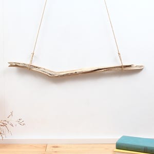 May include: A long, light-coloured piece of driftwood suspended horizontally by two thin, tan-coloured ropes. The wood features a natural, weathered texture with visible grain and organic curves, hanging from a single point above the centre.