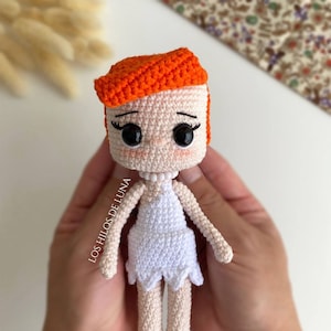 Puede incluir: Una muñeca de crochet con pelo naranja, vestido blanco y piel marrón. La muñeca lleva un collar de perlas y tiene grandes ojos negros. La muñeca está siendo sostenida en las manos de una persona.