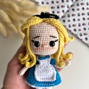 May include: A crocheted doll of Alice from Alice in Wonderland. The doll is wearing a blue dress with a white apron and has blonde hair with a black headband. The doll is holding a small white rabbit.