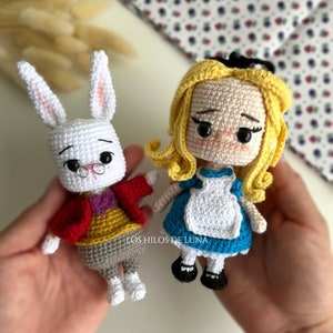 Puede incluir: Dos mu&ntilde;ecos de ganchillo: un conejo blanco con una chaqueta roja y una ni&ntilde;a rubia con un vestido azul. El conejo lleva gafas y una pajarita morada. La ni&ntilde;a tiene un lazo negro en el pelo. Los mu&ntilde;ecos est&aacute;n hechos a mano.
