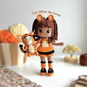 May include: A handmade crochet doll and a small tiger toy. The doll has brown hair, orange and black clothing, and tiger ear headbands. The tiger toy is orange and black. The text "Los Hilos de Luna" is visible.