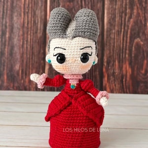 May include: A crocheted doll with gray hair, a red dress, and a blue jewel on her chest. The doll is standing on a wooden surface. The text "LOS HILOS DE LUNA" is visible on the doll's dress.