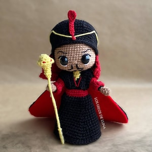 May include: A crocheted amigurumi doll of Jafar from Disney's Aladdin. The doll is wearing a black robe with a red collar and a gold crown. He is holding a golden staff with a snake head on top. The doll is standing on a brown surface.