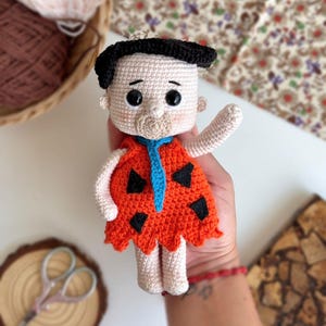 May include: A crocheted plush toy of a cartoon character wearing an orange and black costume. The toy has black hair, a white face, and large black eyes.