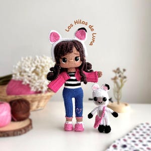 Op de afbeelding: Twee gehaakte poppen, een grote en een kleine, staan naast elkaar. De grotere pop draagt een roze jas, een blauwe broek en een witte hoed met roze oren. De kleinere pop is een witte beer met een roze tas. De tekst "Los Hilos de Luna" is zichtbaar.