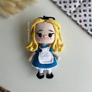 Puede incluir: Mu&ntilde;eca de ganchillo de Alicia en el Pa&iacute;s de las Maravillas, con cabello rubio, vestido azul y delantal blanco. Zapatos negros y lazo negro. El texto "Los Hilos de Luna" es visible. Producto artesanal.