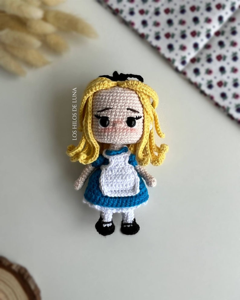 Puede incluir: Mu&ntilde;eca de ganchillo de Alicia en el Pa&iacute;s de las Maravillas, con cabello rubio, vestido azul y delantal blanco. Zapatos negros y lazo negro. El texto "Los Hilos de Luna" es visible. Producto artesanal.