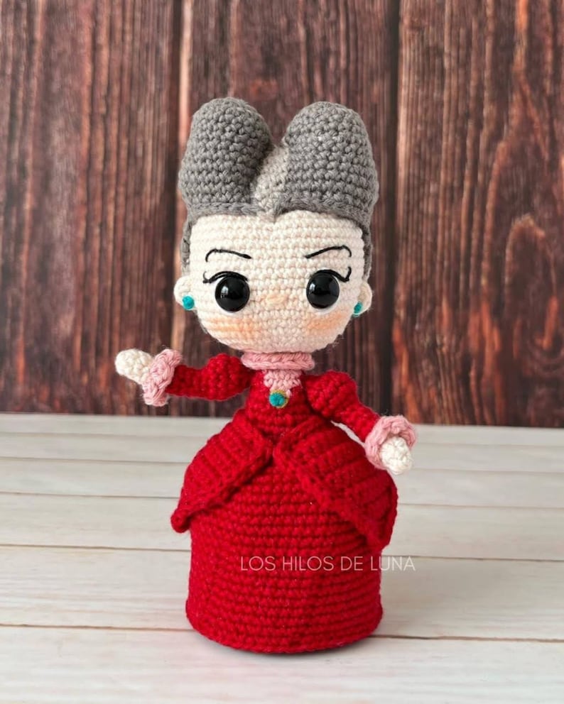 Pode incluir: Uma boneca de croch&ecirc; com cabelo cinza, um vestido vermelho e uma joia azul no peito. A boneca est&aacute; em p&eacute; sobre uma superf&iacute;cie de madeira. O texto "LOS HILOS DE LUNA" &eacute; vis&iacute;vel no vestido da boneca.