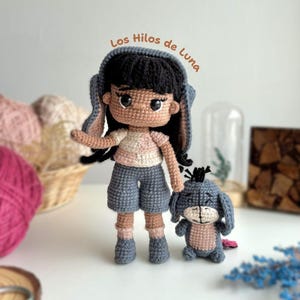 Könnte beinhalten: Eine gehäkelte Amigurumi-Puppe mit brauner Haut, schwarzen Haaren und einem blauen und rosa Outfit. Die Puppe hält einen gehäkelten blauen und rosa Eeyore aus Winnie Puuh. Der Text "Los Hilos de Luna" ist im Hintergrund geschrieben.