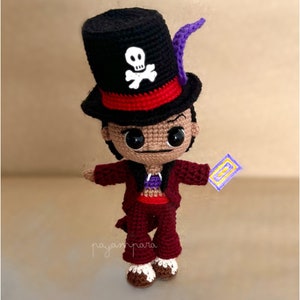 以下が含まれることがあります： 骸骨と交差した骨のデザイン、紫色の羽根、赤と黒のジャケット、赤いズボンをつけたキャラクターの編みぐるみぬいぐるみ。おもちゃは、10と書かれた紫と黄色のカードを持っています。